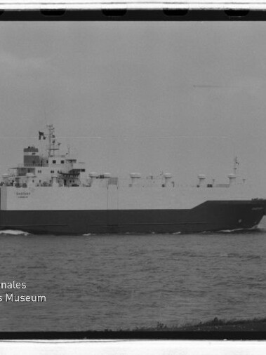 RoRo-Frachtschiff mit hellem Oberrumpf und dunklem Unterrumpf fährt auf einem Fluss; Museumslogo links unten.