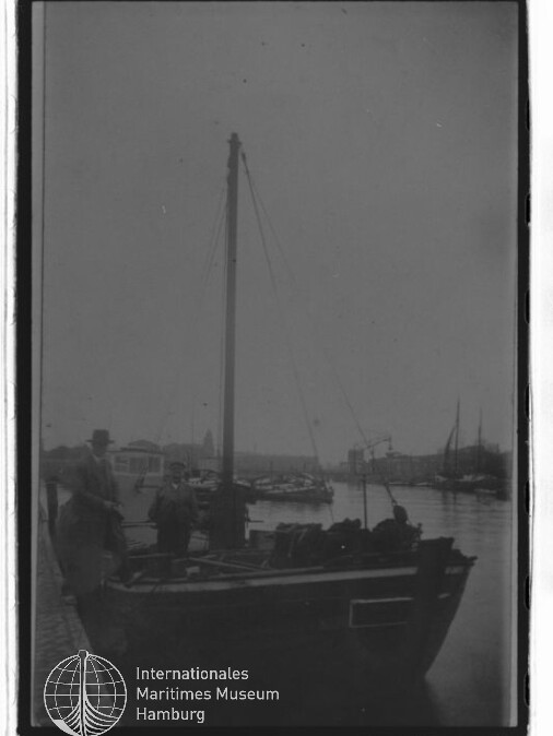 Kleines Segelboot am Kai mit zwei Personen, düstere Hafenkulisse und sichtbares Logo Internationales Maritimes Museum Hamburg
