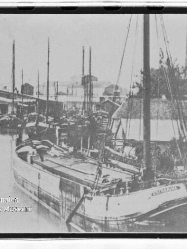 Schwarzweißaufnahme eines Kanals mit mehreren festgemachten Segelkähnen und hohen Masten neben Lagerhäusern.