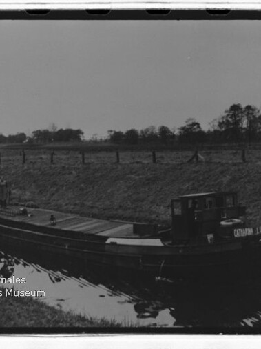 Schwarzweißaufnahme eines langen Lastkahns auf einem Kanal mit kleinem Steuerhaus und flachem Deck, Ufer mit Bäumen im Hintergrund.