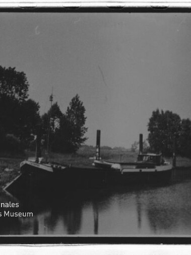 Schwarzweißfoto eines am Ufer vertäuten Lastkahns vor Baumreihe, ruhige Wasserfläche mit Spiegelung und Museumslogo links unten.