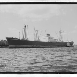 Großes Frachtschiff am Kai mit Hafenkränen im Hintergrund, zwei Schlepper auf dem Wasser und Museumslogo links unten.