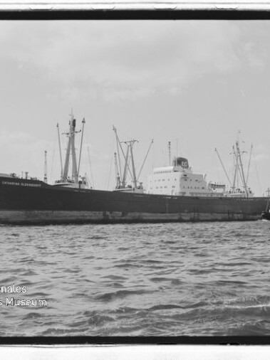 Großes Frachtschiff am Kai mit Hafenkränen im Hintergrund, zwei Schlepper auf dem Wasser und Museumslogo links unten.