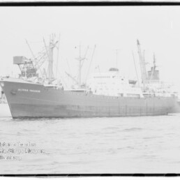 Großes Frachtschiff am Kai eines Industriehafens mit Ladekränen im Hintergrund und Wasser im Vordergrund.