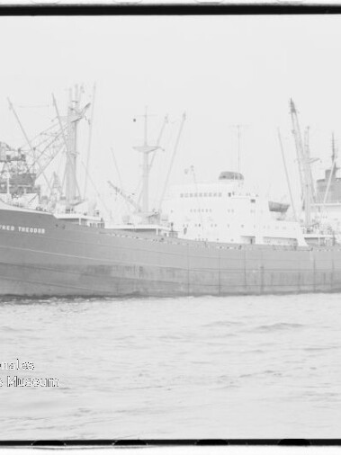 Großes Frachtschiff am Kai eines Industriehafens mit Ladekränen im Hintergrund und Wasser im Vordergrund.