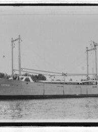 Schwarzweißaufnahme eines Frachtschiffs CATRUN mit zwei Masten und Ladebäumen, auf ruhigem Wasser vor Uferkulisse.