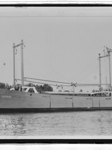 Schwarzweißaufnahme eines Frachtschiffs CATRUN mit zwei Masten und Ladebäumen, auf ruhigem Wasser vor Uferkulisse.