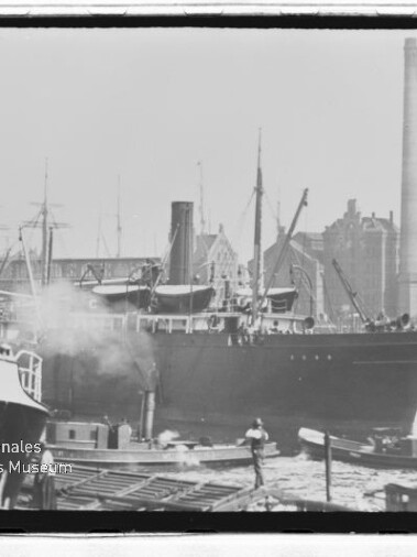 Großes Dampfschiff am Hafenpier, von kleineren Booten umgeben, mit rauchendem Schornstein und Industriegebäuden im Hintergrund.