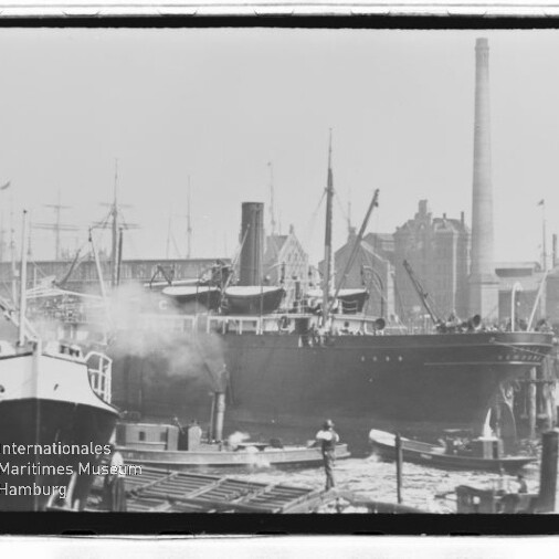 Großes Dampfschiff am Hafenpier, von kleineren Booten umgeben, mit rauchendem Schornstein und Industriegebäuden im Hintergrund.