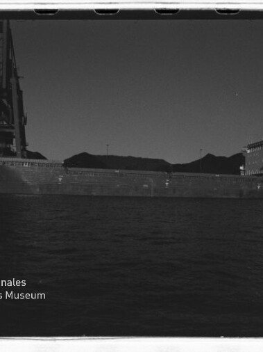 Massives Frachtschiff an einem Hafen mit großem Kran und Lagerhügeln im Hintergrund, Schwarzweißfoto mit Museumswasserzeichen