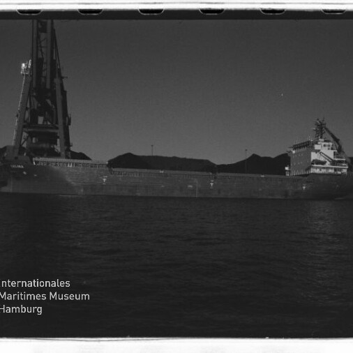 Massives Frachtschiff an einem Hafen mit großem Kran und Lagerhügeln im Hintergrund, Schwarzweißfoto mit Museumswasserzeichen