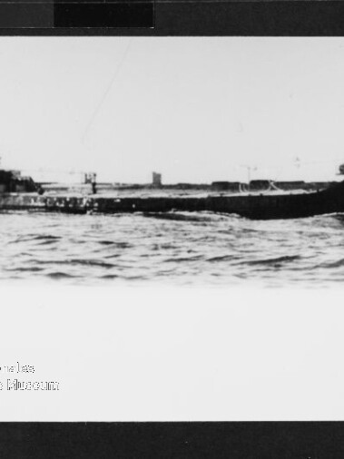 Schwarzweißfoto eines flachen Frachtschiffs mit niedrigem Aufbau auf ruhiger See; Stempel unten links.