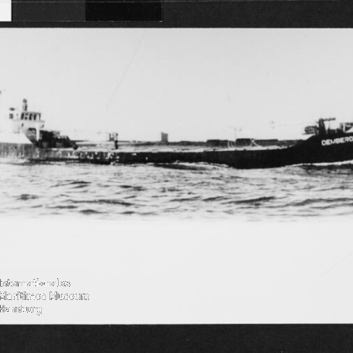 Schwarzweißfoto eines flachen Frachtschiffs mit niedrigem Aufbau auf ruhiger See; Stempel unten links.