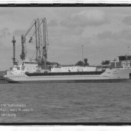 Schwarzweißfoto eines Frachtschiffs am Kai mit Hafenkränen im Hintergrund und Wasser im Vordergrund; Museumswasserzeichen.