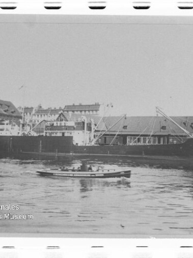 Schwarzweißfoto eines Frachtschiffs am Kai, kleines Boot im Vordergrund und Hafenbauten im Hintergrund; Museumsstempel links unten.
