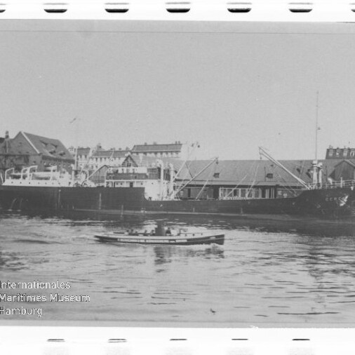 Schwarzweißfoto eines Frachtschiffs am Kai, kleines Boot im Vordergrund und Hafenbauten im Hintergrund; Museumsstempel links unten.