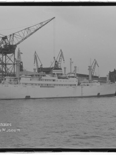 Schwarzweißfoto eines großen weißen Schiffes im Dock vor mehreren Hafenkränen; Museumslogo links unten.