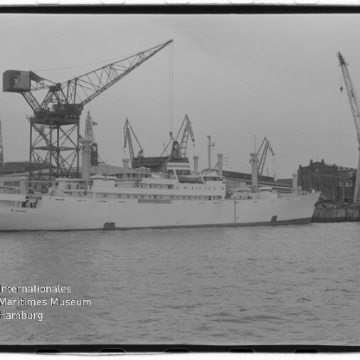 Schwarzweißfoto eines großen weißen Schiffes im Dock vor mehreren Hafenkränen; Museumslogo links unten.