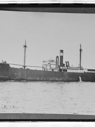 Frachtschiff mit hohem Schornstein und zwei Masten, Name am Bug sichtbar, fährt auf ruhiger See.