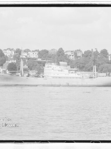 Graustufiges Frachtschiff auf ruhiger See vor einem von Häusern gesäumten Ufer, Wasserzeichen unten links