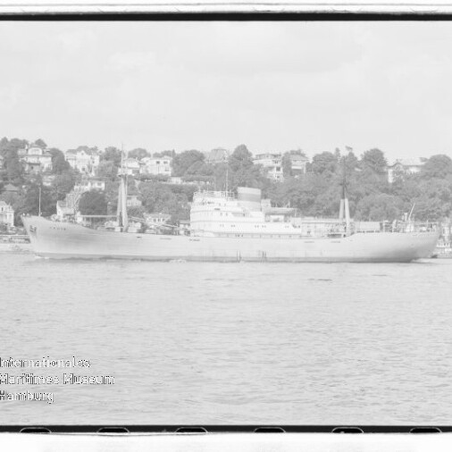 Graustufiges Frachtschiff auf ruhiger See vor einem von Häusern gesäumten Ufer, Wasserzeichen unten links