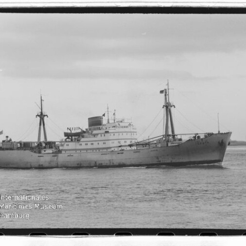 Graustufiges Frachtschiff mit zwei Masten und Aufbauten vor flacher Küste, mit Wasserzeichen Maritimes Museum Hamburg.