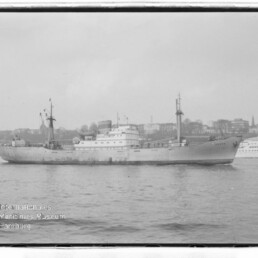 Schwarzweißaufnahme eines Frachtschiffs auf ruhigem Wasser vor einer Stadtkulisse; links unten Wasserzeichen Internationales Maritimes Museum Hamburg