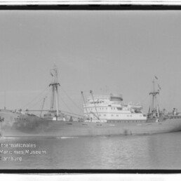 Schwarzweißfoto eines großen Frachtschiffs mit Aufbauten und Ladegeschirr im Hafen, Museumslogo unten links.