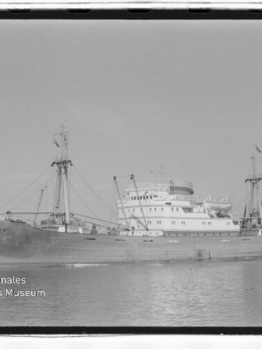 Schwarzweißfoto eines großen Frachtschiffs mit Aufbauten und Ladegeschirr im Hafen, Museumslogo unten links.