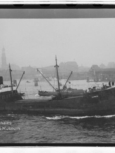 Frachtschiff fährt in Hafen mit nebliger Stadtkulisse und Kirchturm im Hintergrund, Museumswasserzeichen unten links.