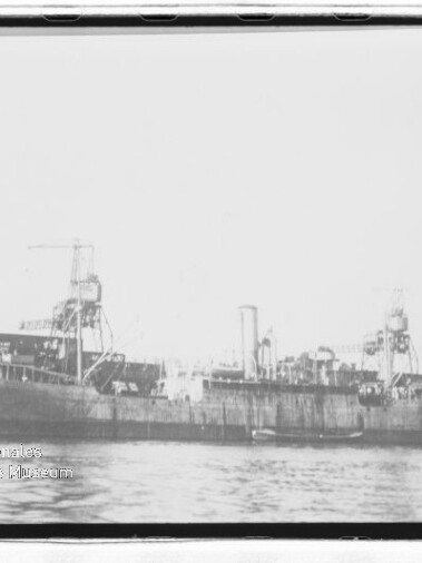 Langes, altes Frachtschiff mit Deckkränen und mittigem Schornstein am Kai, ruhiges Wasser im Vordergrund.