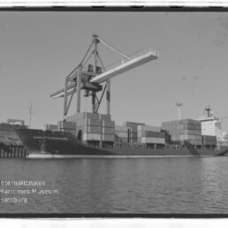 Containerschiff mit gestapelten Containern an einem großen Hafenkran, Wasser im Vordergrund und Wasserzeichen Internationales Maritimes Museum Hamburg