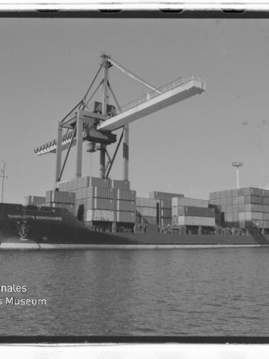Containerschiff mit gestapelten Containern an einem großen Hafenkran, Wasser im Vordergrund und Wasserzeichen Internationales Maritimes Museum Hamburg