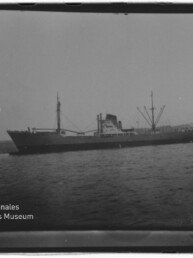 Frachtschiff mit Ladekränen und Schlepper im Hafen, Wasser im Vordergrund; Stempel Internationales Maritimes Museum Hamburg.