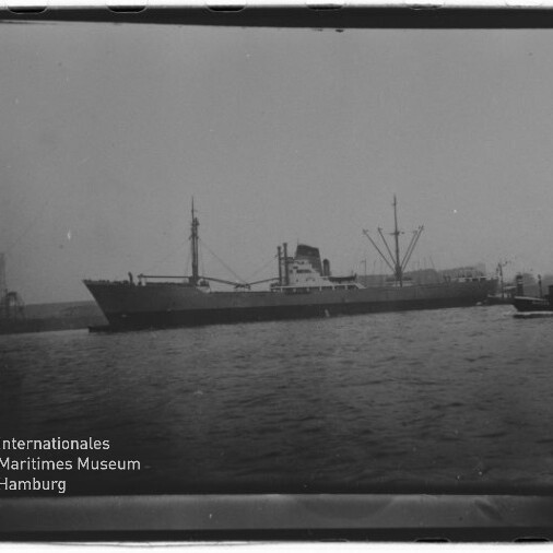 Frachtschiff mit Ladekränen und Schlepper im Hafen, Wasser im Vordergrund; Stempel Internationales Maritimes Museum Hamburg.