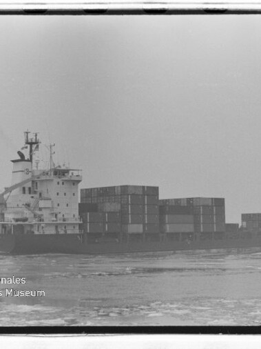 Containerschiff mit gestapelten Containern fährt bei nebligem Himmel über leicht eisbedecktes Meer; Museumslogo unten links.