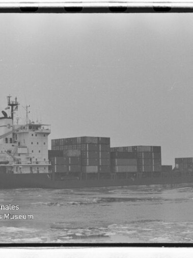 Frachtschiff mit aufgetürmten Containern fährt auf ruhiger See; schwarz-weiß-Aufnahme mit Wasserzeichen des Internationalen Maritimen Museums Hamburg.