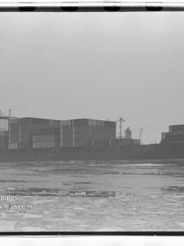 Containerschiff mit gestapelten Containern auf teils zugefrorenem Meer in Nebel, Wasserzeichen Int. Maritimes Museum Hamburg.
