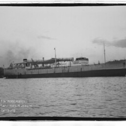 Schwarzweißaufnahme eines großen Dampfschiffs längsseits vor Hafen, ruhige See; Wasserzeichen Internationales Maritimes Museum Hamburg.