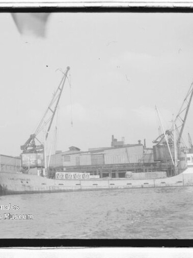 Frachtschiff am Industriekaianleger neben Hafenkränen; schwarzweißaufnahme mit Wasserzeichen des Maritimen Museums Hamburg.