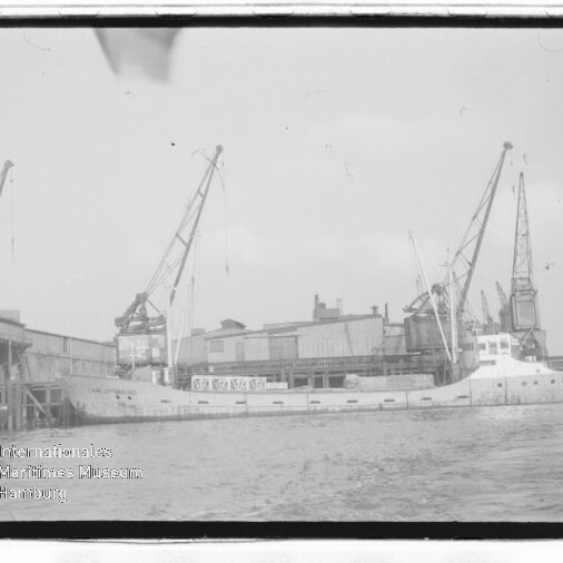Frachtschiff am Industriekaianleger neben Hafenkränen; schwarzweißaufnahme mit Wasserzeichen des Maritimen Museums Hamburg.