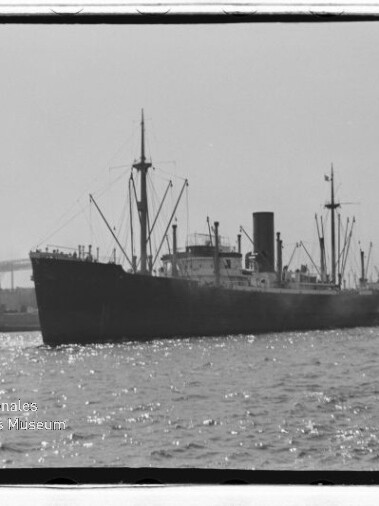 Schwarzweiß-Foto eines Frachtschiffs mit Schornstein und Ladebäumen im Hafen, im Hintergrund Hafenkräne und Museumswasserzeichen.