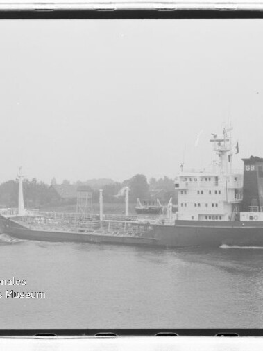 Schwarzweißfoto eines Tankschiffs auf ruhigem Wasser vor einem baumbestandenen Ufer, Museum-Logo unten links.