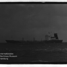 Frachtschiff mit Aufbauten achtern auf rauer See unter dunklem Himmel; links unten Wasserzeichen Maritimes Museum Hamburg.