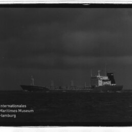 Schwarzweißfoto eines Tankschiffs auf rauer See unter dunklen Wolken, Museumslogo unten links