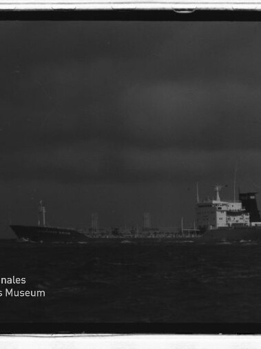 Schwarzweißfoto eines Tankschiffs auf rauer See unter dunklen Wolken, Museumslogo unten links