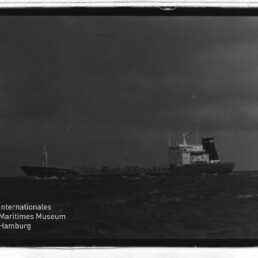 Schwarzweiß-Frachtschiff mit Heckaufbau und Schornstein auf See; unten links Logo Maritimes Museum Hamburg.