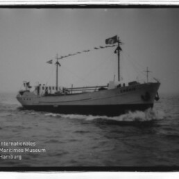Schwarzweißfoto eines Frachtschiffs mit Flaggen auf See; Schriftzug Internationales Maritimes Museum sichtbar.
