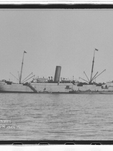 Dampfschiff mit Schornstein und zwei Masten, mit Fahnen auf ruhigem Wasser vor Küste; kleiner Schlepper daneben.