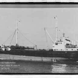 Schwarzweißaufnahme eines Frachtschiffs mit zwei Masten und Aufbauten, fahrend auf ruhigem Wasser; Museumslogo unten links.